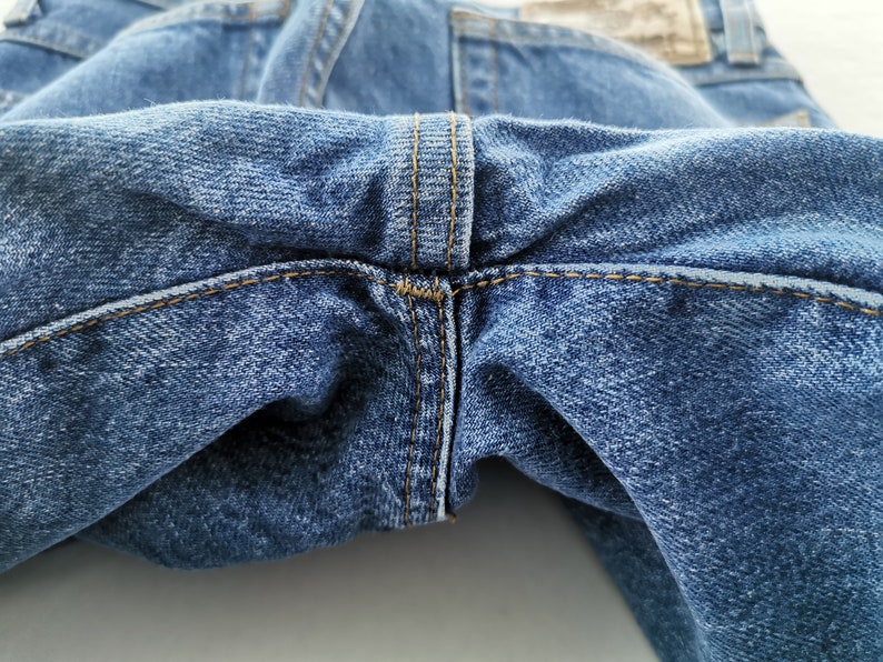 May include: Close-up of blue denim jeans, showcasing the stitching details at the crotch and waistband. The denim has a classic, worn look with visible texture and subtle fading. The stitching is in a contrasting tan color.