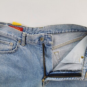 May include: Light blue denim jeans with a partially unzipped fly. The jeans have a classic five-pocket design, a button closure, and a red tag. The denim has a faded, worn look.
