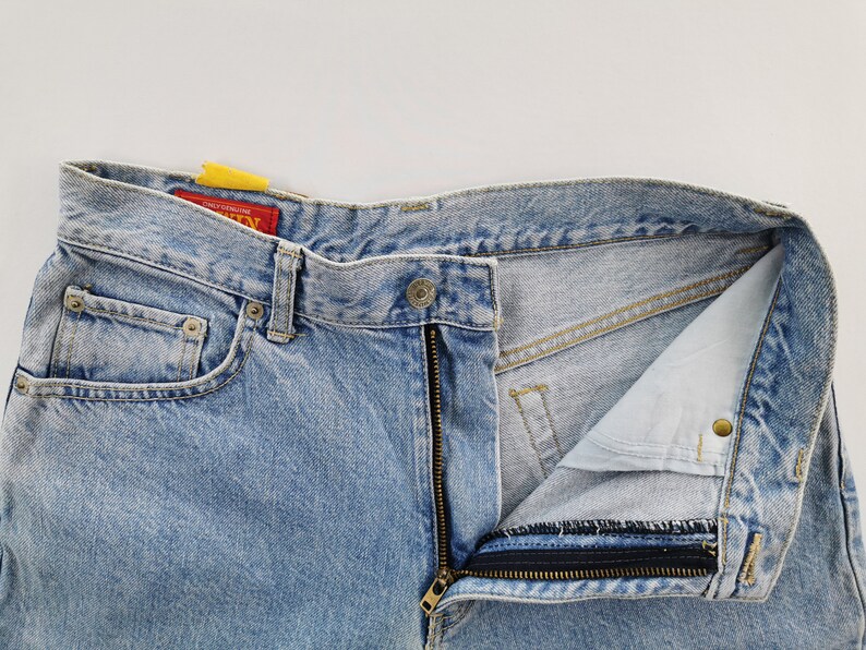May include: Light blue denim jeans with a partially unzipped fly. The jeans have a classic five-pocket design, a button closure, and a red tag. The denim has a faded, worn look.