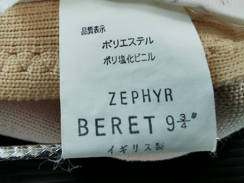 Puede incluir: Primer plano de una etiqueta de boina. La etiqueta dice "ZEPHYR BERET 9 3/4" e incluye texto en japon&eacute;s. La boina es de color beige claro. La etiqueta tambi&eacute;n incluye el texto "Made in England."