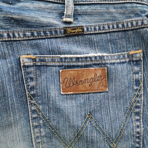May include: Close-up of blue denim jeans with a Wrangler brand leather patch on the back pocket. The jeans have a classic five-pocket design with yellow stitching and a small Wrangler label above the pocket.