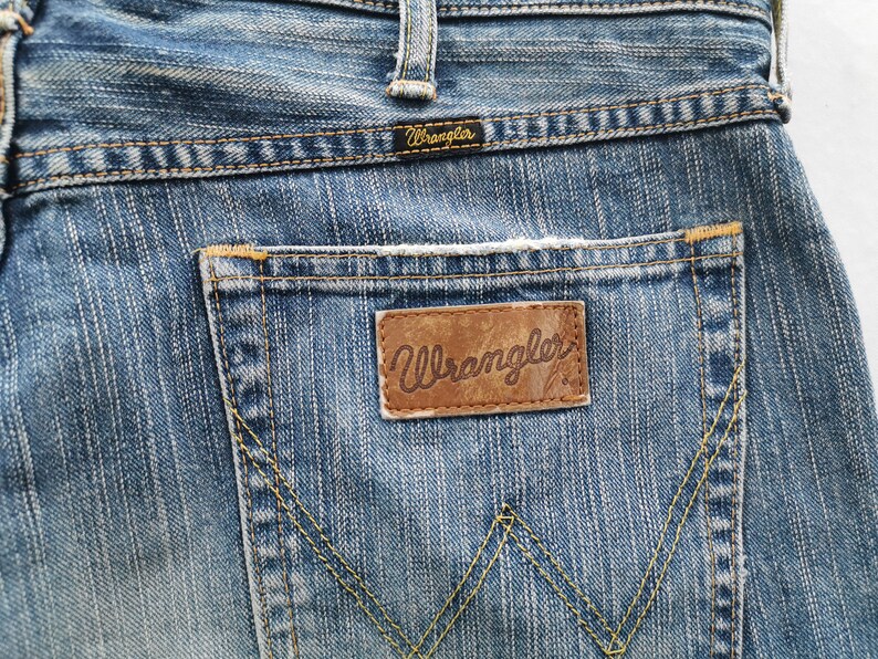 May include: Close-up of blue denim jeans with a Wrangler brand leather patch on the back pocket. The jeans have a classic five-pocket design with yellow stitching and a small Wrangler label above the pocket.