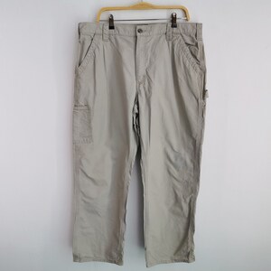 May include: Khaki-colored work pants hanging on a wooden hanger against a white wall. The pants have multiple pockets, including a cargo pocket on the side. The pants are made of a durable material and have a relaxed fit.