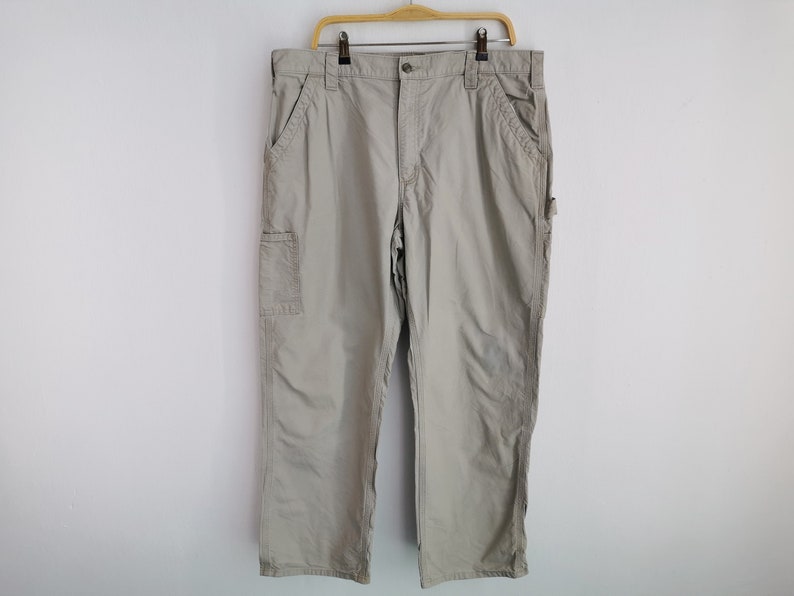 May include: Khaki-colored work pants hanging on a wooden hanger against a white wall. The pants have multiple pockets, including a cargo pocket on the side. The pants are made of a durable material and have a relaxed fit.