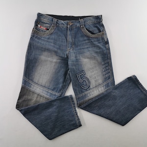 May include: Blue denim jeans featuring a faded wash, a prominent embroidered number 5 on the left leg, and a rectangular patch with text. The pants include a button fly, front pockets, and dark grey fabric panels integrated into the lower legs.