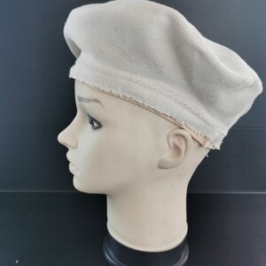 Puede incluir: Boina beige de tejido de punto. La boina se muestra en una cabeza de maniqu&iacute;, mostrando su forma y textura. El borde de la boina tiene un acabado en bruto. El fondo es una superficie oscura.