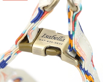 May include: A pet harness featuring a cream-colored fabric strap with a multicolored pattern of blue, orange, and red accents. The harness includes a metallic bronze-toned side-release buckle engraved with the name Isabella and the number 647-635-0832.