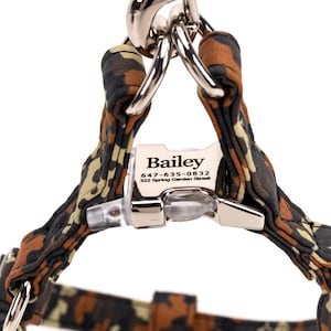 May include: A dog harness with a camouflage pattern in shades of brown, black, and tan. The harness features a silver metal clasp and a white tag with the name "Bailey" and contact information.