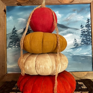 May include: A stack of four fabric pumpkins in red, orange, white, and red. The pumpkins are arranged in a pyramid shape and tied together with twine. The pumpkins are sitting on a brown and white patterned surface.