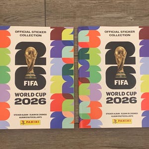 May include: Two official Panini sticker albums for the FIFA World Cup 2026. The covers feature a gold trophy, the FIFA logo, and the text "WORLD CUP 2026" against a colorful background with the text "OFFICIAL STICKER COLLECTION".