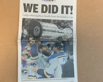 Pode incluir: Uma manchete de jornal diz "WE DID IT!" com o subtítulo "Long-suffering Blues finally hoist the Stanley Cup". O jornal é o St. Louis Post-Dispatch, uma edição final da Copa Stanley. A imagem mostra um grupo de jogadores de hóquei celebrando com o troféu da Copa Stanley.