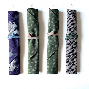 Peut inclure: Quatre rouleaux enveloppés de tissu différents avec les numéros 1 à 4 imprimés au-dessus de chaque rouleau. Les rouleaux sont enveloppés de motifs différents, dont un rouleau violet avec un motif de dragon, un rouleau vert avec un motif floral, un rouleau vert avec un motif géométrique et un rouleau marron avec un motif géométrique.