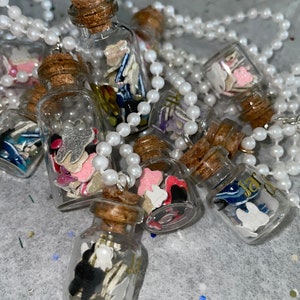 Puede incluir: Un collar con varias botellas pequeñas de vidrio llenas de confeti y abalorios coloridos. Las botellas están unidas a una cadena de perlas blancas.