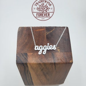 May include: A silver necklace with the word "aggies" in a cursive font. The necklace is displayed on a wooden block. The image also includes a circular logo with the text "Traditions Forever".