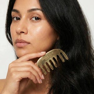 May include: A gold-toned metal hair comb with a curved top edge and wide, rounded teeth. The surface of the comb features the engraved text Herbal Edge and Naturally Yours. The comb is held against dark hair, showcasing its metallic finish.