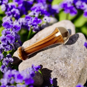May include: A wooden bottle stopper with a gold-colored top rests on a gray stone, surrounded by purple flowers. The stopper has a bulbous end and a cylindrical body with visible wood grain.