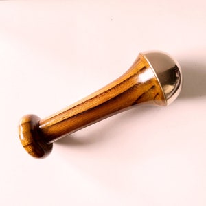 May include: A wooden pestle with a polished, cylindrical handle and a rounded base. The top features a shiny, silver-colored metal cap. The wood grain is visible, showcasing a warm, brown color.