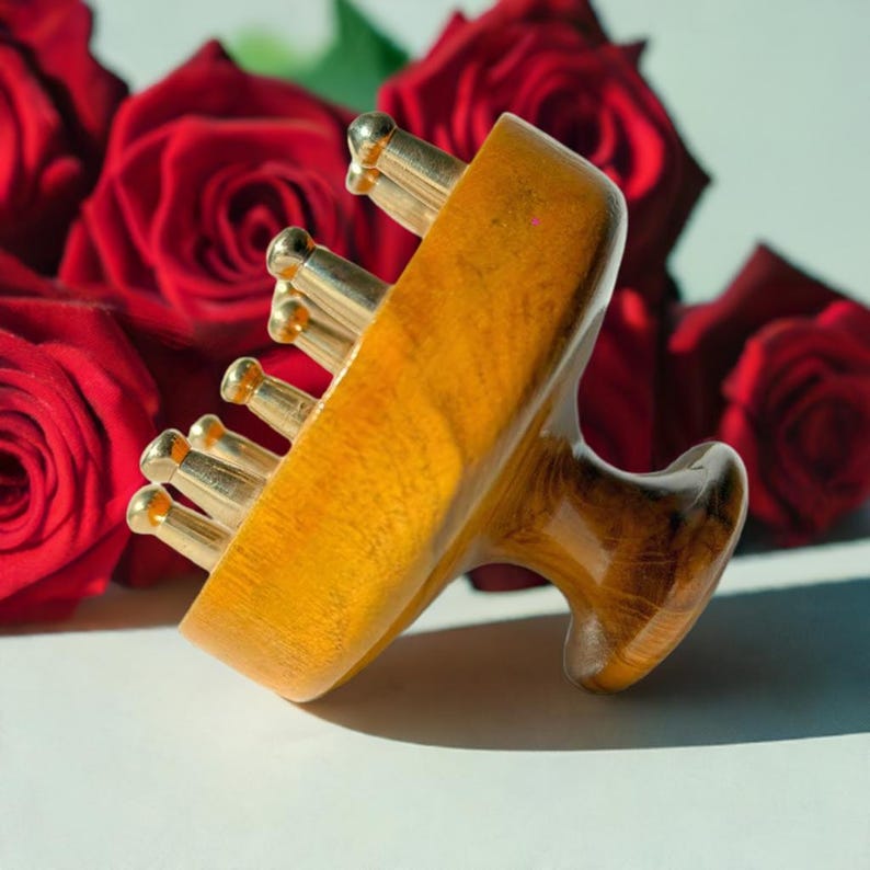 May include: A wooden massage tool with gold-colored metal rollers. The tool has a handle and is made of light brown wood. Red roses are in the background.