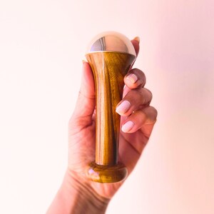May include: A wooden massage tool with a gold-colored, rounded top. The tool has a cylindrical wooden handle and a wider base. The wood grain is visible, and the tool is held against a light pink background.