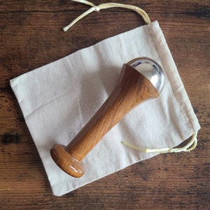 May include: A wooden facial massage tool with a silver sphere at the top, resting on a white cloth bag. The tool has a polished, light brown finish. The bag is made of a natural-colored fabric. The background is a wooden surface.