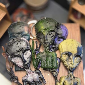 May include: A collection of alien-shaped candles in various colors, including green, purple, black, and gold. The candles are arranged on a wooden surface, with some holding up peace signs.