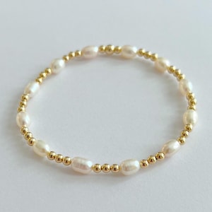 May include: A delicate bracelet featuring creamy white oval pearls interspersed with small, round gold beads. The bracelet is strung on an elastic band, creating a flexible and elegant design. The jewelry is set against a plain white background.