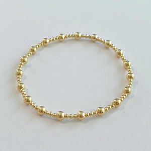 May include: A gold beaded bracelet. The bracelet is made of small, round, gold-colored beads strung together to form a circular shape. The beads are evenly spaced and create a delicate, elegant design. The bracelet is shown on a white surface.
