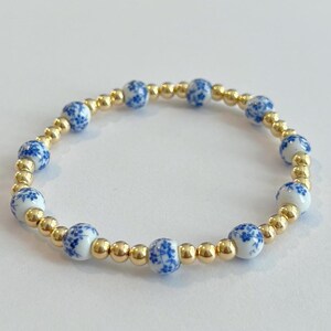 May include: A beaded bracelet featuring alternating gold-colored beads and white ceramic beads with blue floral patterns. The bracelet is circular and rests on a white surface. The jewelry is likely for fashion or decorative purposes.