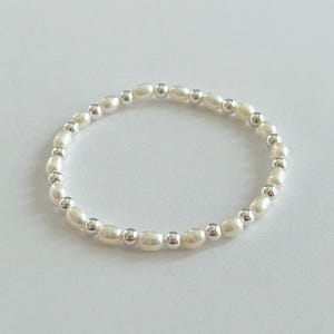 May include: A delicate bracelet featuring alternating white oval pearls and small silver-colored beads. The bracelet is circular and rests on a white surface, showcasing its simple elegance. This jewelry piece is suitable for everyday wear.