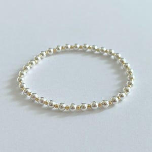Puede incluir: Una pulsera de cuentas con cuentas plateadas y doradas alternas. La pulsera está hecha de pequeñas cuentas redondas ensartadas, creando un accesorio delicado y elegante. El diseño es simple y versátil, adecuado para el uso diario.