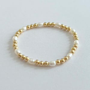 May include: A delicate beaded ring featuring alternating gold-colored and white pearl beads. The ring is circular and sits on a white surface. The beads are small and evenly spaced, creating a simple yet elegant design.