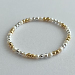 May include: A delicate bracelet featuring a mix of small, round beads. The bracelet alternates between gold, silver, and white beads, creating a subtle contrast. The beads are strung together to form a flexible, circular shape, suitable for wrist wear.