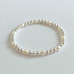 May include: A delicate ring featuring alternating white oval pearls and small silver beads. The ring is set against a white background, highlighting the contrast between the bright pearls and the metallic beads. A simple and elegant piece of jewelry.