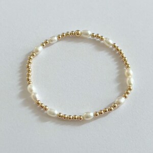 May include: A delicate bracelet featuring alternating white oval pearls and small gold-colored beads. The bracelet is strung on an elastic band, creating a flexible and elegant design. The jewelry is set against a white background.