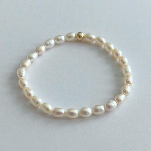 May include: A delicate bracelet featuring small, oval-shaped white pearls strung together. A single gold-colored bead is centered on the bracelet. The jewelry is displayed on a white surface, highlighting the natural luster of the pearls.