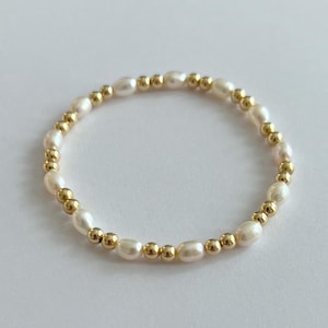 May include: A delicate bracelet featuring creamy white oval pearls and small gold-colored beads. The pearls are strung together with the gold beads interspersed throughout the design. The bracelet is shown on a white background.