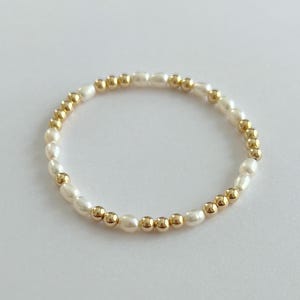 May include: A delicate ring featuring alternating white oval pearls and small gold-colored beads. The ring is set against a plain white background, highlighting the jewelry's elegant design. This piece is suitable for everyday wear or special occasions.