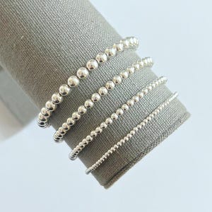 Puede incluir: Cuatro pulseras de cuentas plateadas de diferentes tamaños se exhiben sobre un cilindro cubierto de tela gris. Las pulseras presentan pequeñas cuentas redondas y brillantes ensartadas. Las cuentas varían en tamaño, creando un efecto en capas. El fondo es gris claro.