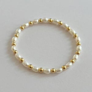 May include: A delicate bracelet featuring white oval pearls and small gold-colored beads. The pearls are strung together with the gold beads evenly spaced throughout the bracelet. The jewelry is displayed on a white surface.