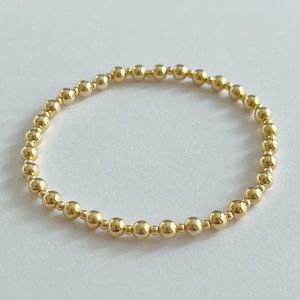 May include: A gold beaded ring. The ring is made of small, round, gold-colored beads strung together to form a continuous circle. The beads have a shiny, metallic finish. The ring is shown against a plain white background.