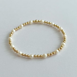 May include: A delicate bracelet featuring alternating white pearls and small gold-colored beads. The bracelet is circular and rests on a white surface, showcasing its elegant design. This jewelry piece is suitable for various occasions.