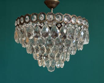 May include: A tiered chandelier featuring multiple clear, teardrop-shaped glass crystals suspended from a circular, antique brass-toned metal frame with a decorative hexagonal border pattern and a central mounting canopy at the top.