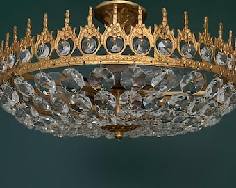May include: A gold-colored chandelier with a crown-like design. The chandelier features clear, faceted crystal pendants and a decorative, ornate frame. The fixture is suspended against a dark teal background, creating a dramatic contrast.
