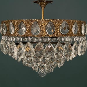 May include: A vintage chandelier with a gold-toned metal frame and clear crystal teardrop-shaped pendants. The upper frame features an ornate design, and the crystals hang in multiple tiers, creating a sparkling effect. The fixture is suspended from a gold-colored ceiling mount.