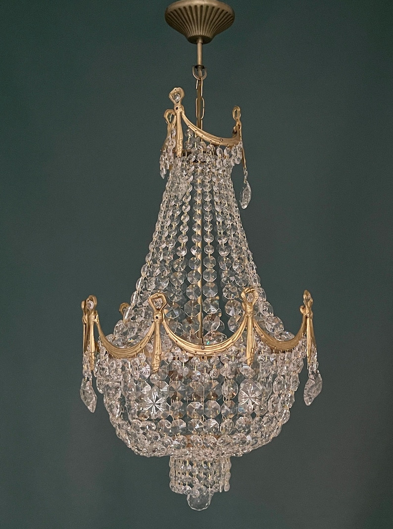 May include: An antique chandelier with a gold-colored frame and clear crystal beads. The chandelier has multiple tiers of crystals and decorative gold accents. It hangs from a matching gold ceiling mount.