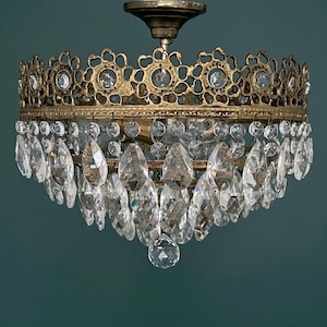 Puede incluir: Lámpara de araña antigua de latón con colgantes de cristal transparente en forma de lágrima. La lámpara de araña tiene un diseño de corona de filigrana decorativa con detalles de cristal transparente. La lámpara está montada en el techo.