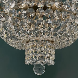 May include: Close-up of a crystal chandelier with multiple tiers of faceted crystals. The chandelier has a gold-colored metal frame and a large, round shape. The crystals reflect light, creating a sparkling effect. The background is a solid teal color.