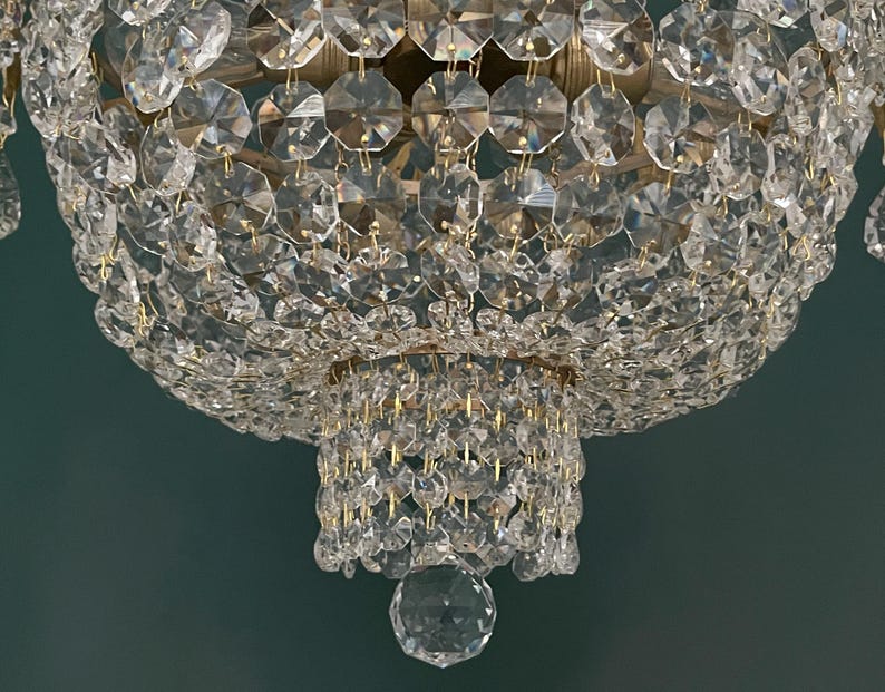 May include: Close-up of a crystal chandelier with multiple tiers of faceted crystals. The chandelier has a gold-colored metal frame and a large, round shape. The crystals reflect light, creating a sparkling effect. The background is a solid teal color.