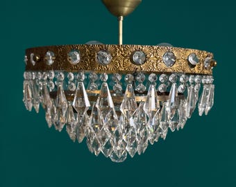 May include: An antique brass chandelier with clear crystal accents. The chandelier features a decorative brass frame with clear crystal embellishments and hanging crystal pendants. The fixture is suspended from a brass ceiling mount, creating a vintage aesthetic.