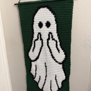 May include: A rectangular crochet wall hanging featuring a white ghost figure on a dark green background. The ghost has black eyes and is depicted with both arms raised, showing middle fingers. The piece is suspended from a thin black rod.